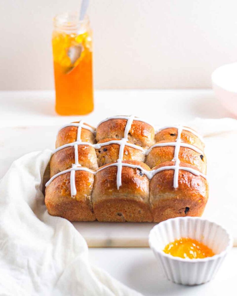 9 hot cross buns with a small bowl of apricot jam and jar in the background