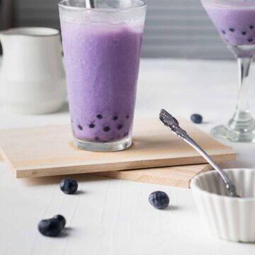 2 glasses filled blueberry boba and have a thick boba straw, with fresh blueberries scattered around it.
