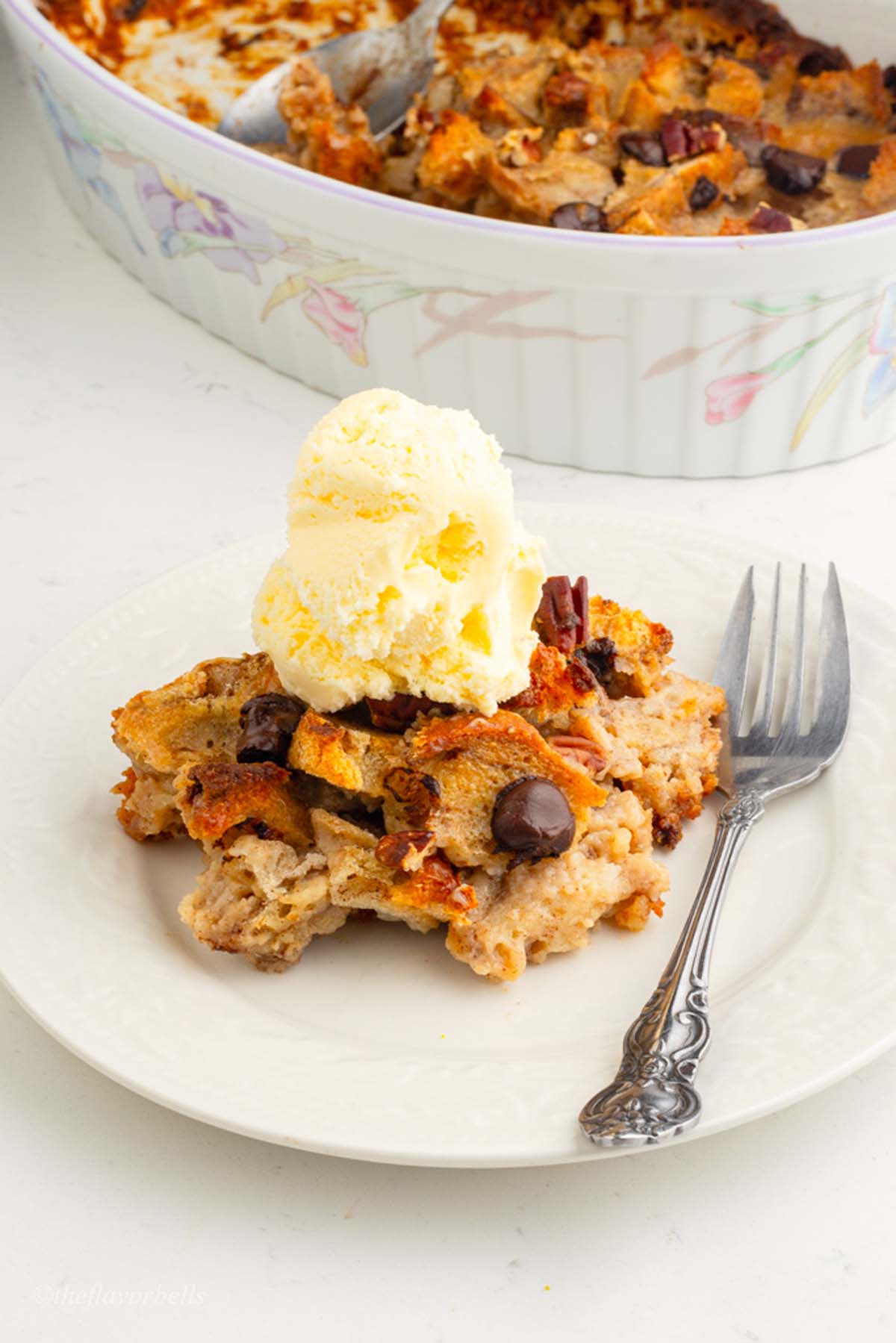 a big scoop of ice cream on the top of bread pudding served in a white plate with casserole of bread pudding in the background