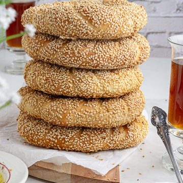5 freshly baked sesame coated simit -Turkish bagels stacked on one another with tea glasses in backdrop and olives and feta cheese in foreground