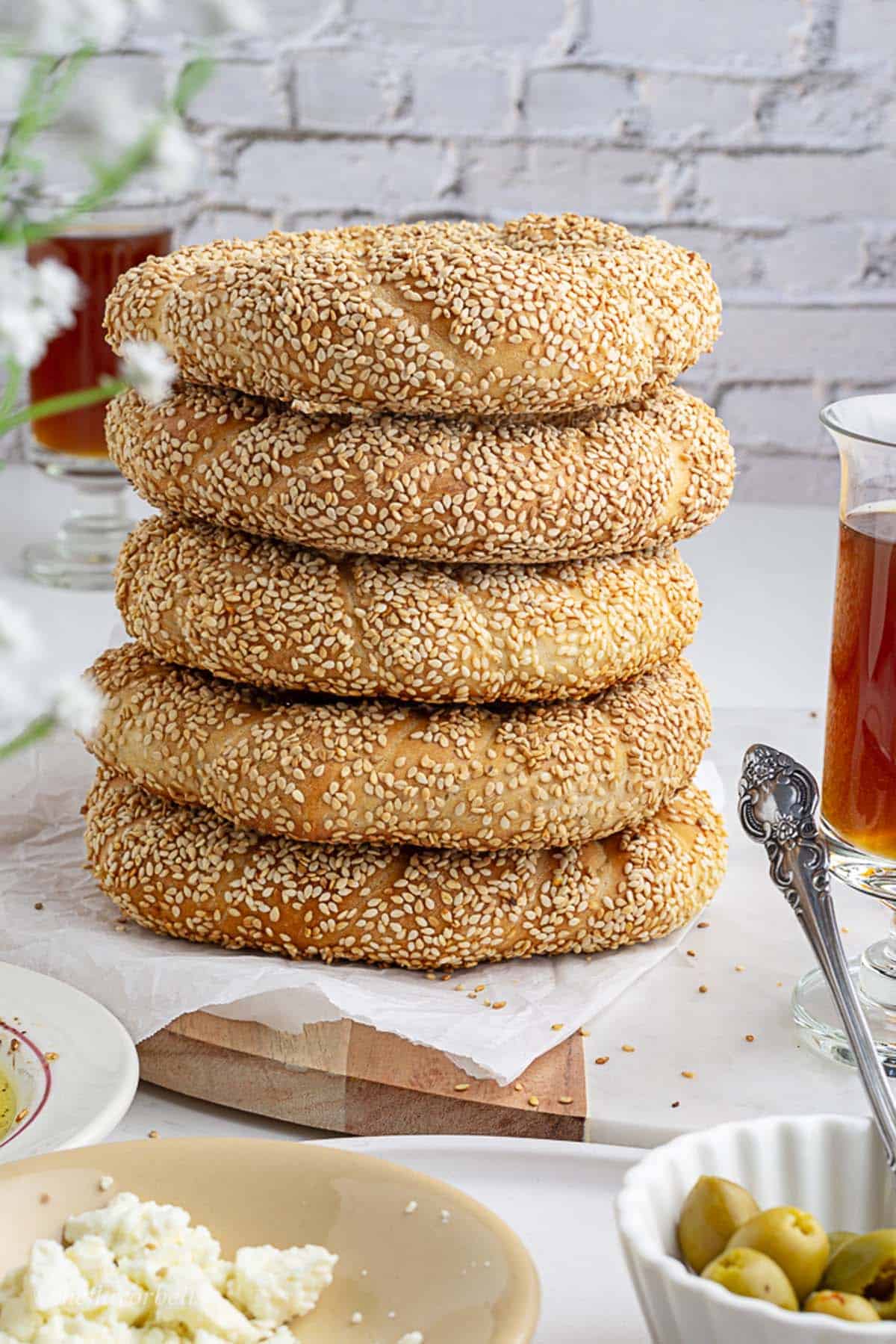 5 freshly baked sesame coated simit -Turkish bagels stacked on one another with tea glasses in backdrop and olives and feta cheese in foreground