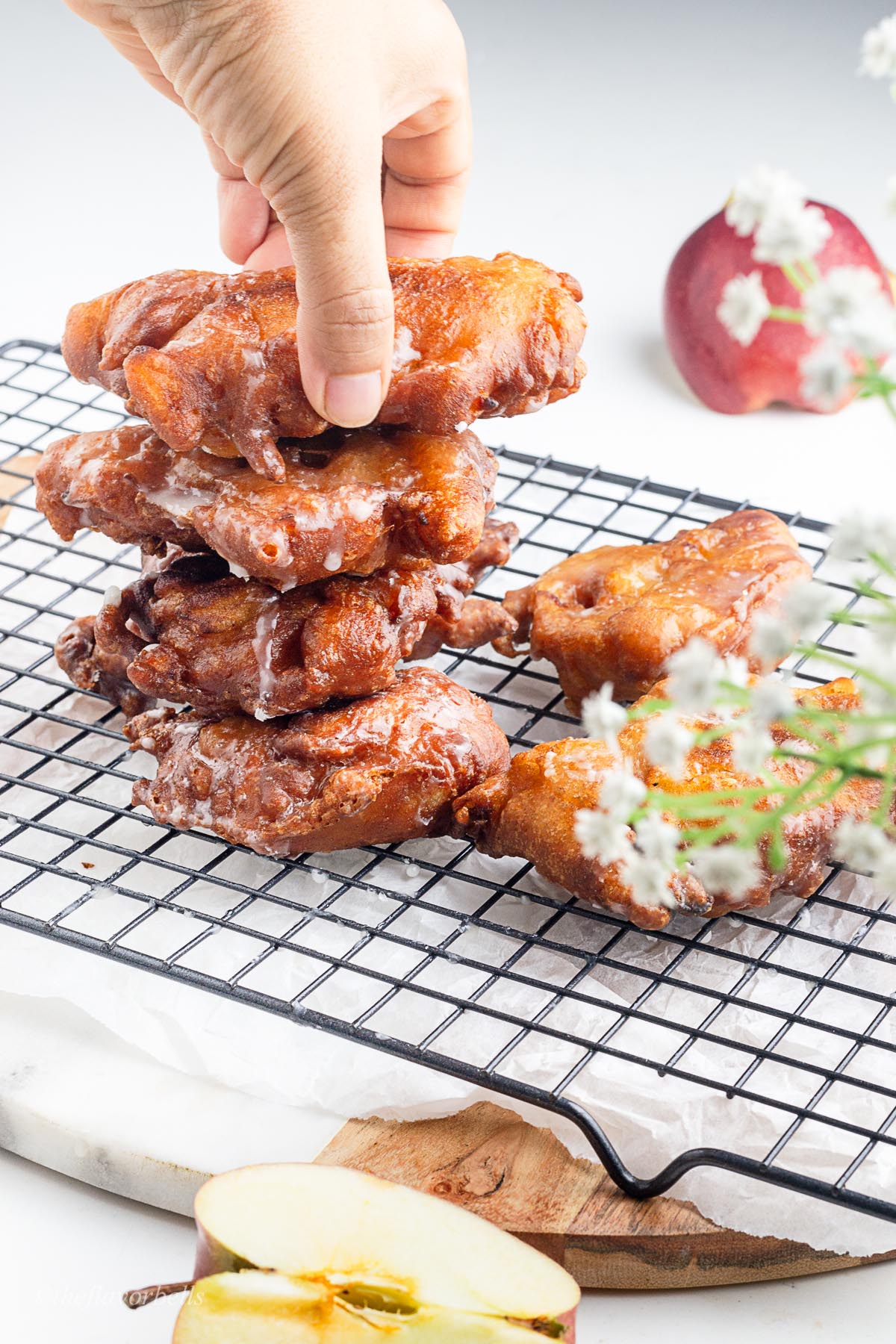 a stack of sourdough apple fritters being placed on a wire rack with some more apple fritters lying around. someone grabbing the fritter from the top of the stack